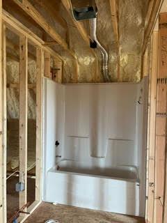 A bathtub set into a framed alcove with spray foam insulation and rough plumbing overhead.