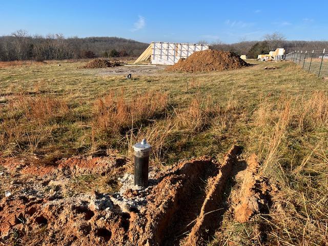 A capped well head standing in a trenched area of red Ozark clay, with open pasture and the house under construction visible in the distance.