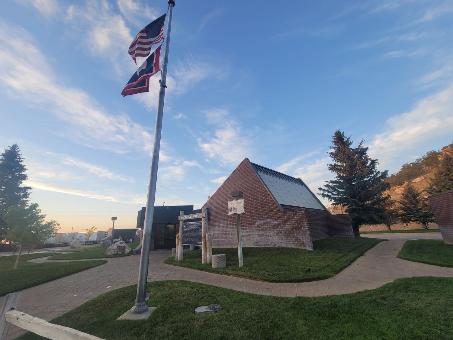 The rest area seen across its lawn at dusk, flagpole in the foreground, earth berming visible around the base of the brick walls.